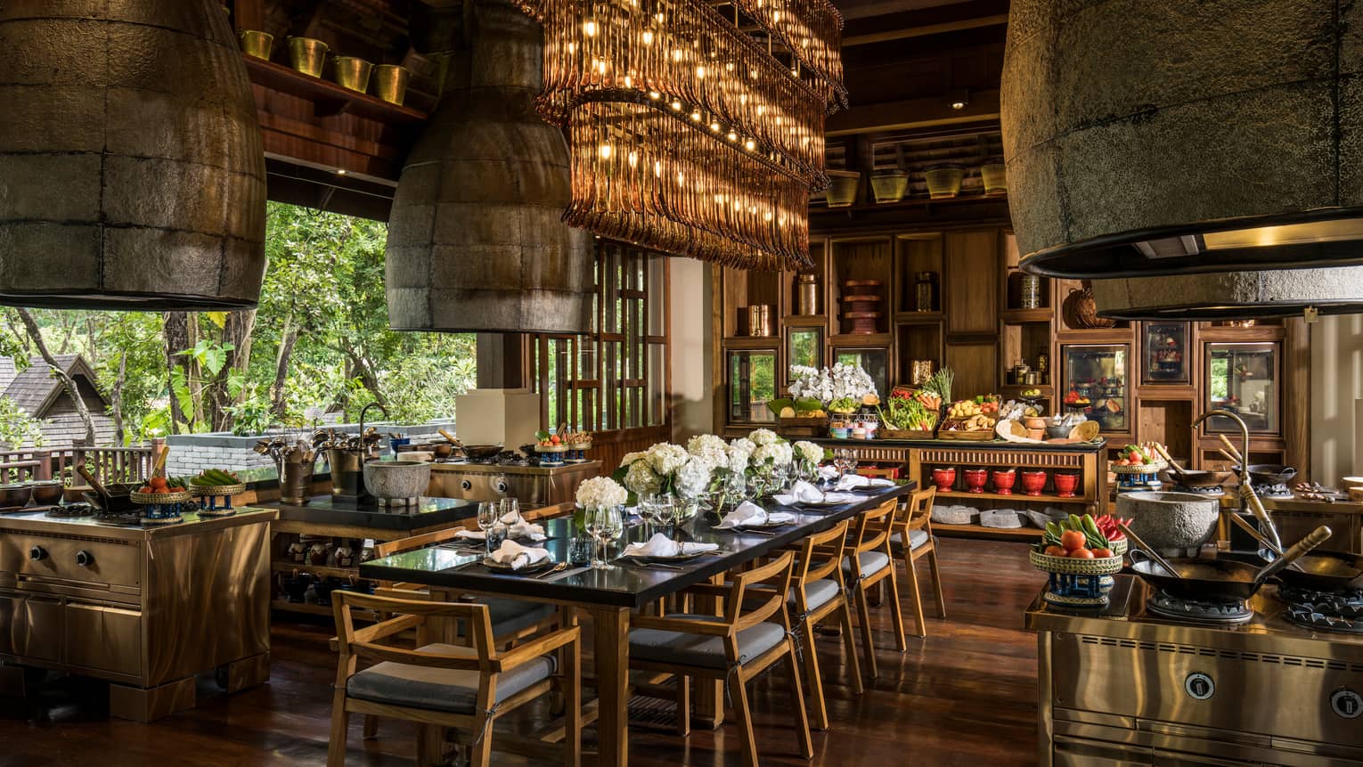 Large rectangular crystal chandelier over long dining table in open kitchen, baskets of vegetables