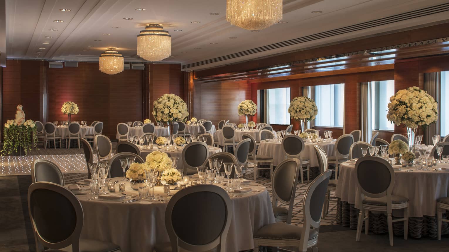 Refined wedding reception area at the Four Seasons London Park Lane in Mayfair, beautifully arranged with silver and white decor. The room features round tables covered in elegant grey tablecloths, each set with pristine white china, crystal glassware, and silver cutlery. Large, lush floral arrangements of white and pale yellow flowers serve as centerpieces, creating an opulent and welcoming atmosphere. The soft, ambient lighting from modern chandeliers and warm wooden accents on the walls add to the room’s luxurious charm.