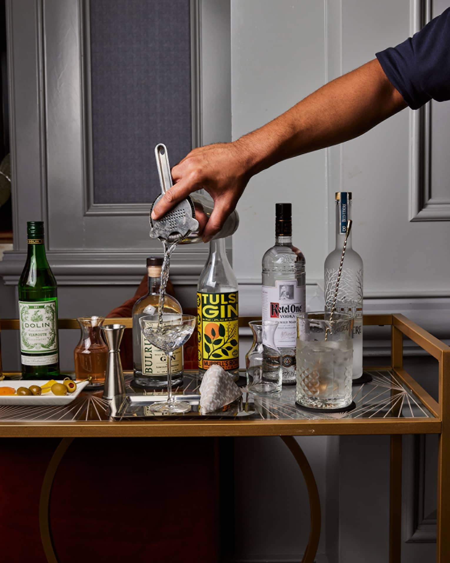 A bartender pours from a shaker into a coupe glass next to bottles of gin, vodka and vermouth, a jigger, carafes and olives.