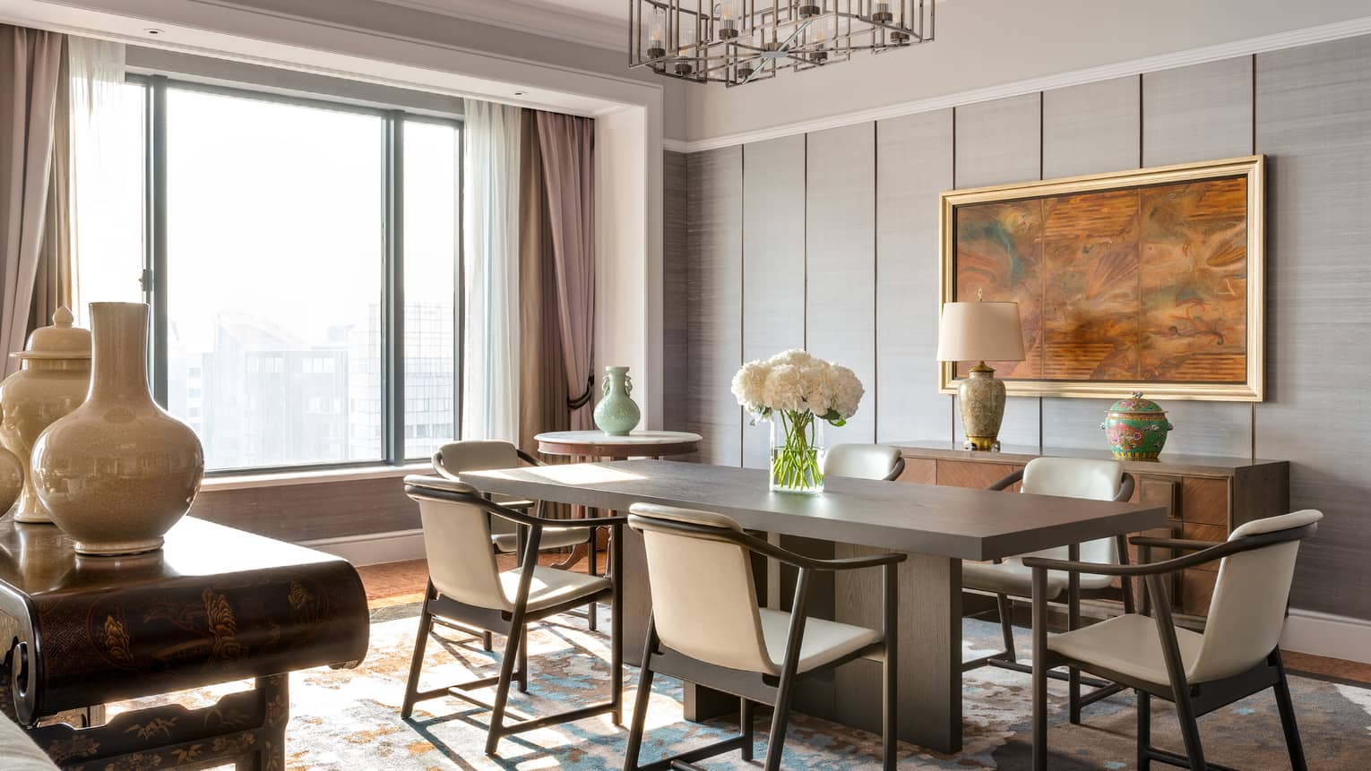 A sophisticated dining room with a rectangular wooden table, cream-coloured chairs, decorative vases and large window. A framed painting and lamp adorn the sideboard.