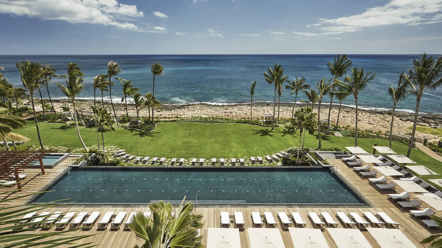 Aerial view of large outdoor swimming pool lined with white deck chairs, lawn, beach