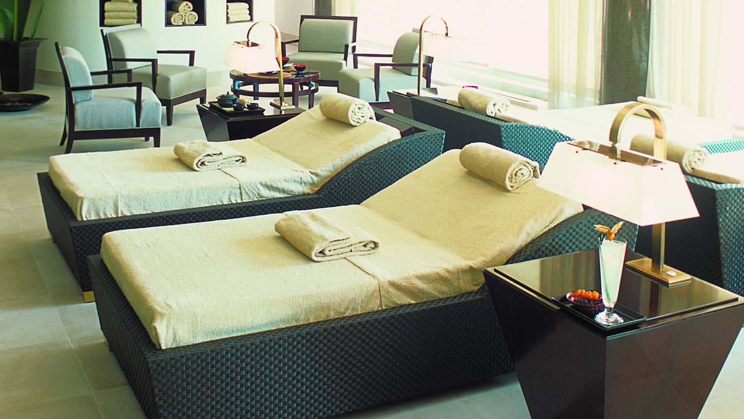 Two large black wicker spa beds with cushions, rolled towels beside table with lamp, seating area in background
