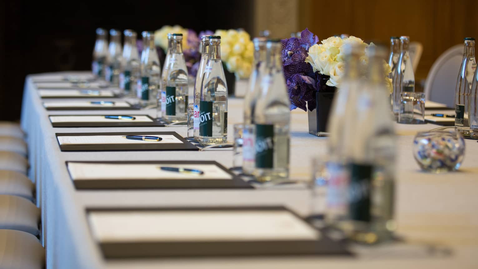 Row of meeting agendas, glass bottles, small vases with flowers on boardroom table