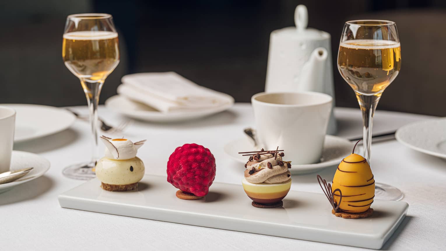 Four colouful, artful pastries lined up on a white rectangular plate served next to two glasses of chmapagne and a mug and pot of tea