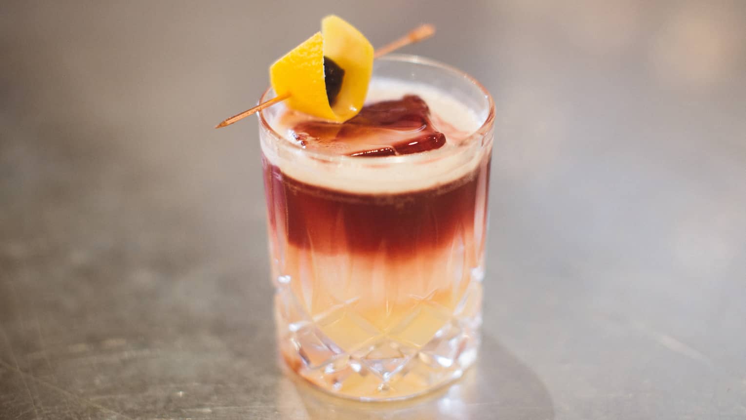 A cocktail with layers of yellow, white and red, topped with a lemon peel