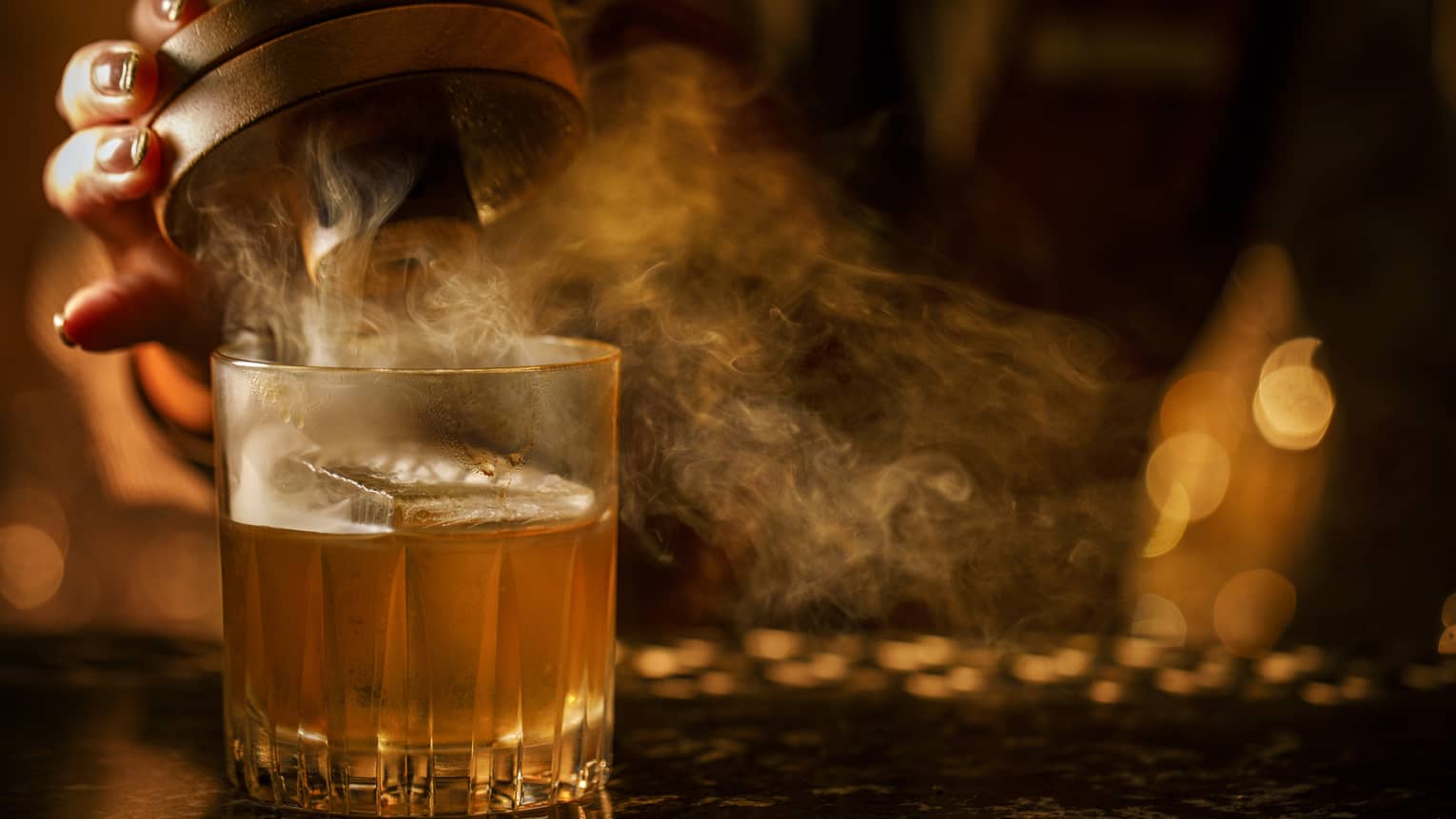 Hand lifts the lid of a smoked Old-Fashioned cocktail in a dimly lit setting