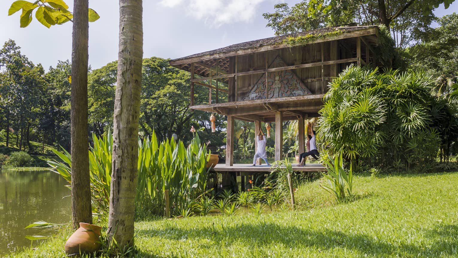 Yogi and two guests kneel with arms raised in yoga posed on pavilion deck near pond