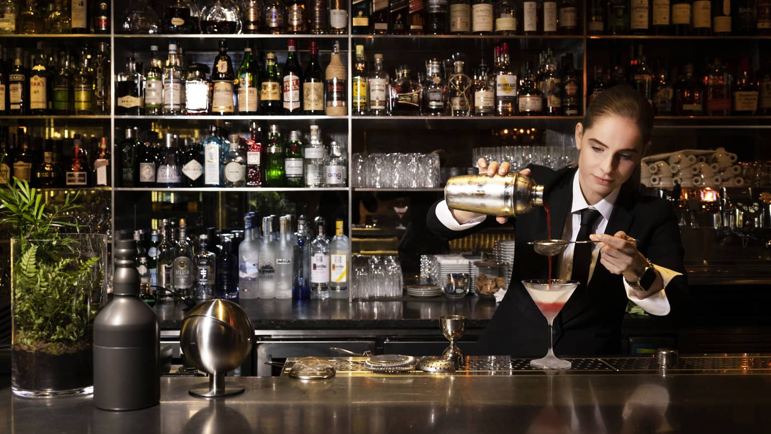 Bartender Silvie wearing a dark suit strains a red cocktail into a Martini glass on bar with stocked shelves behind