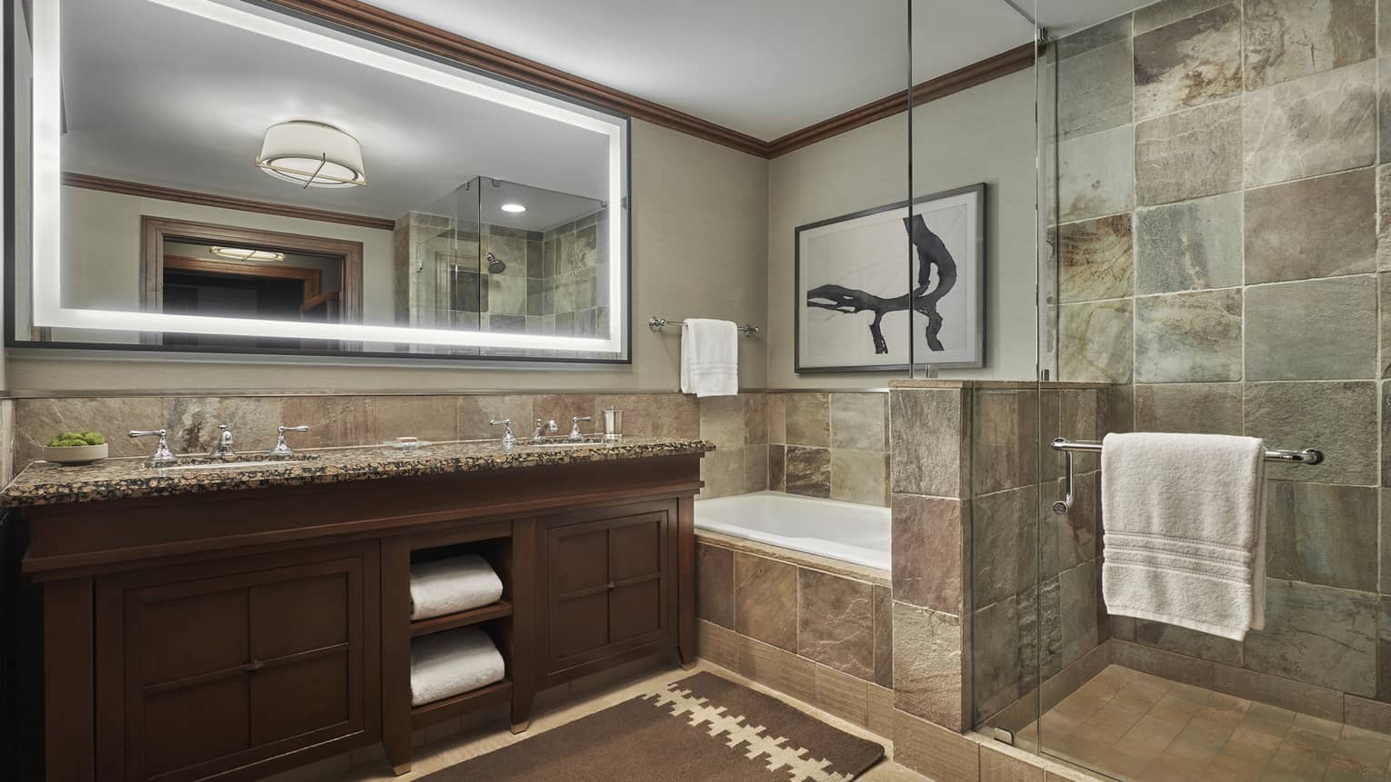 A tiled bathroom, with a double vanity, large lighted mirror and a shower and tub separated by a glass partition