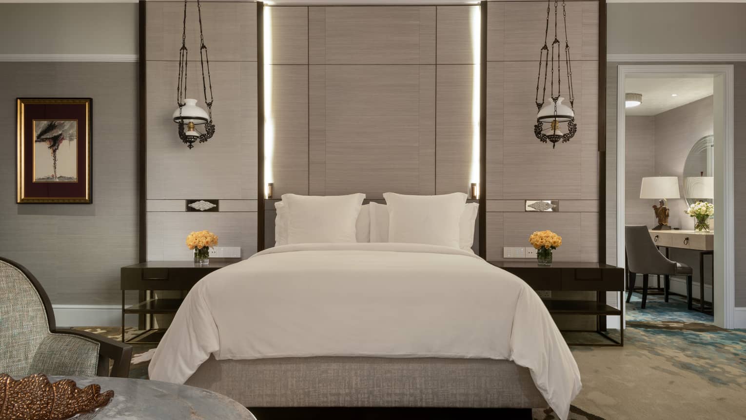 A stylish bedroom with a large bed adorned in white linens, flanked by modern hanging lamps. Nightstands hold vases of yellow roses, and a vanity is visible in the adjacent room.