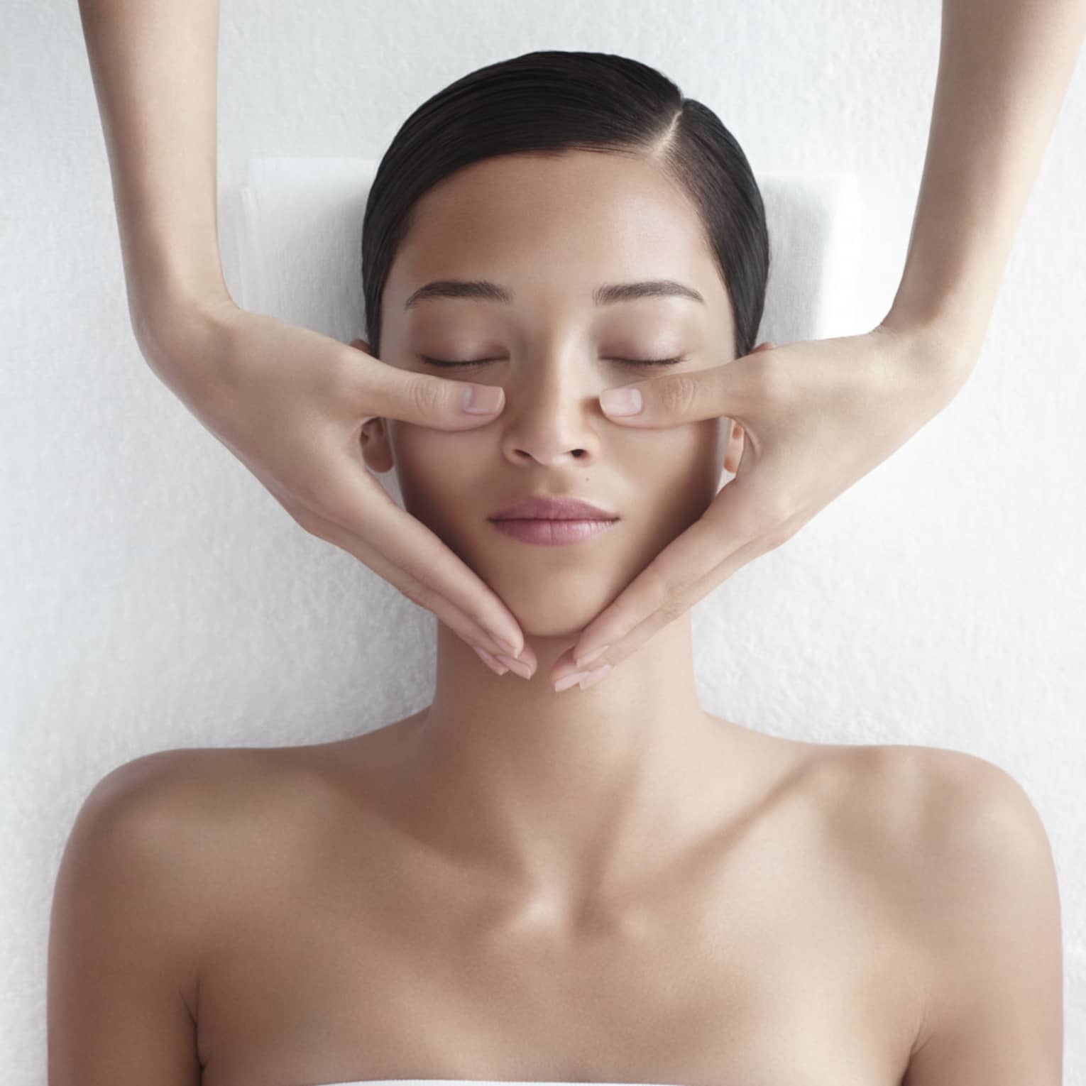 Woman in white bathrobe lies on massage table with eyes closed, masseuse rests hands on her cheeks