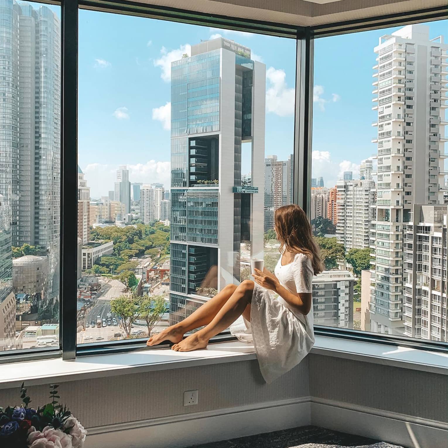 Woman lounges in hotel room corner window overlooking city