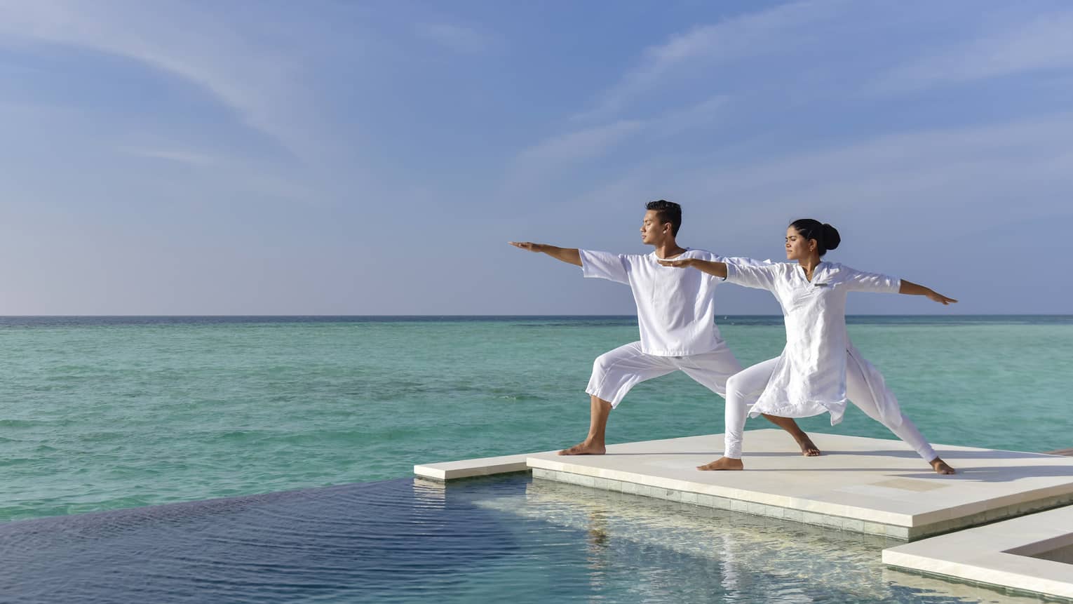 A couple dressed in all white does yoga on a white dock over the sea