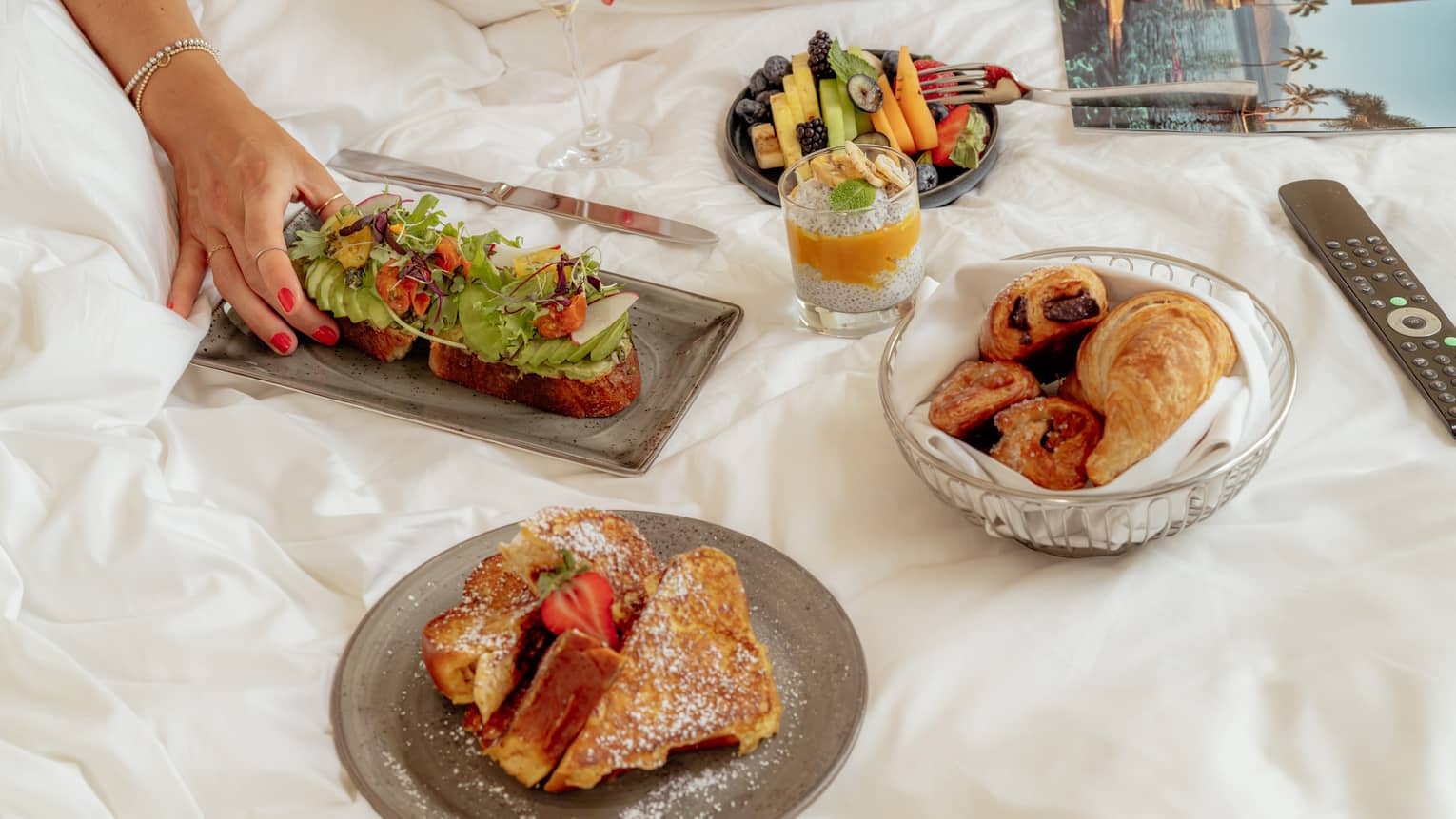 A few small plates of food items like pastries and French toast are arranged on a bed, next to a magazine and remote control. A person's hand reaches for one of the food items.