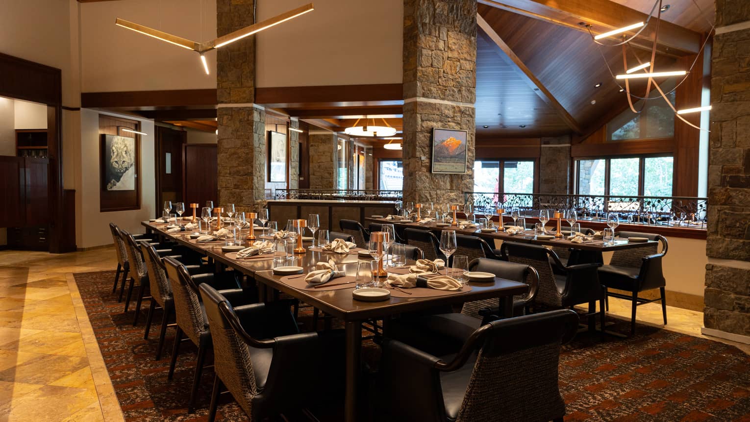 A warm interior dining space with windows on one wall, wood floors, long dining tables set for a meal, chairs, and large three-bladed ceiling fans suspended from the high ceiling