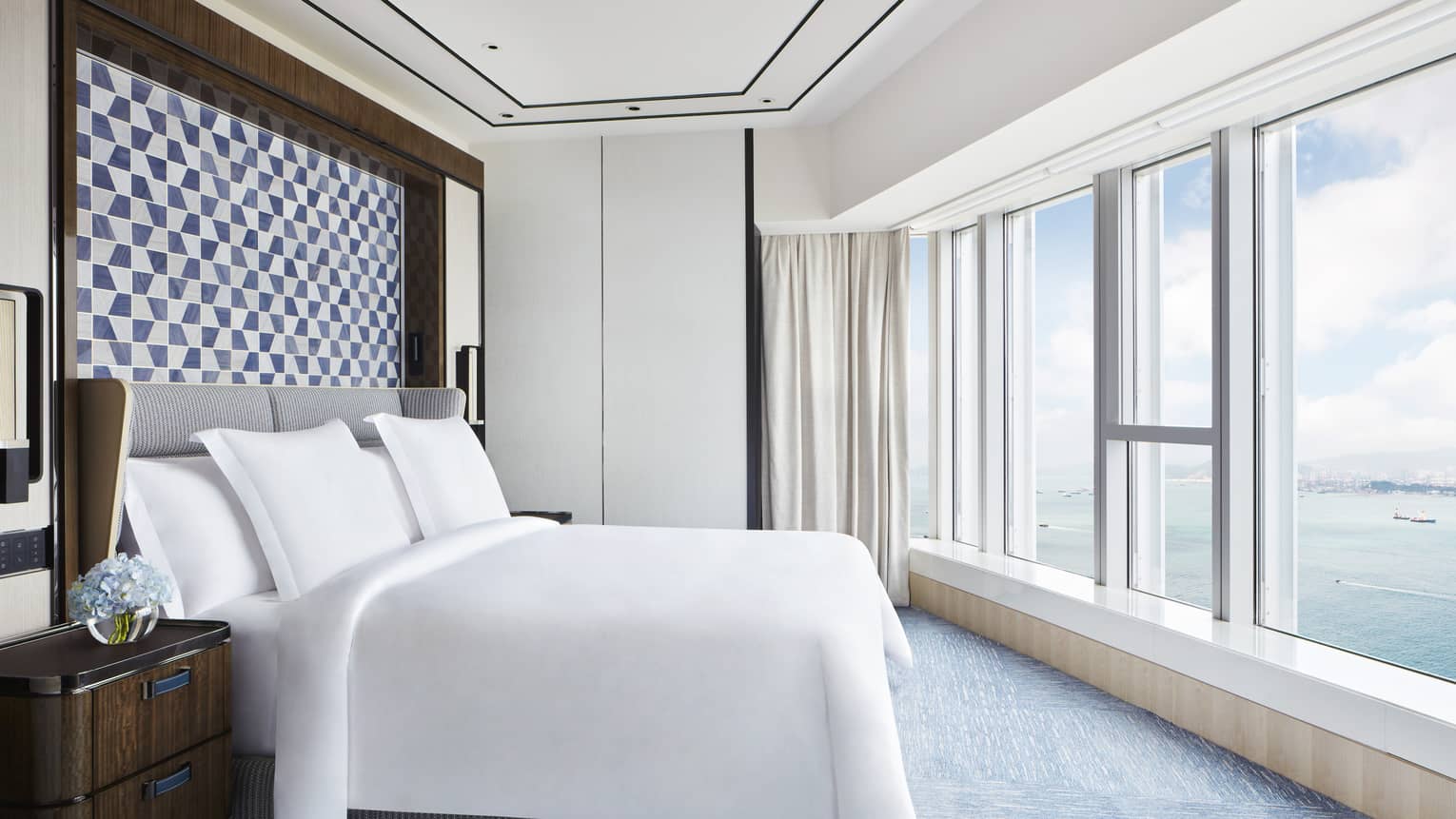 Bright hotel bedroom with large bed, blue geometric headboard, floor-to-ceiling windows and ocean view.