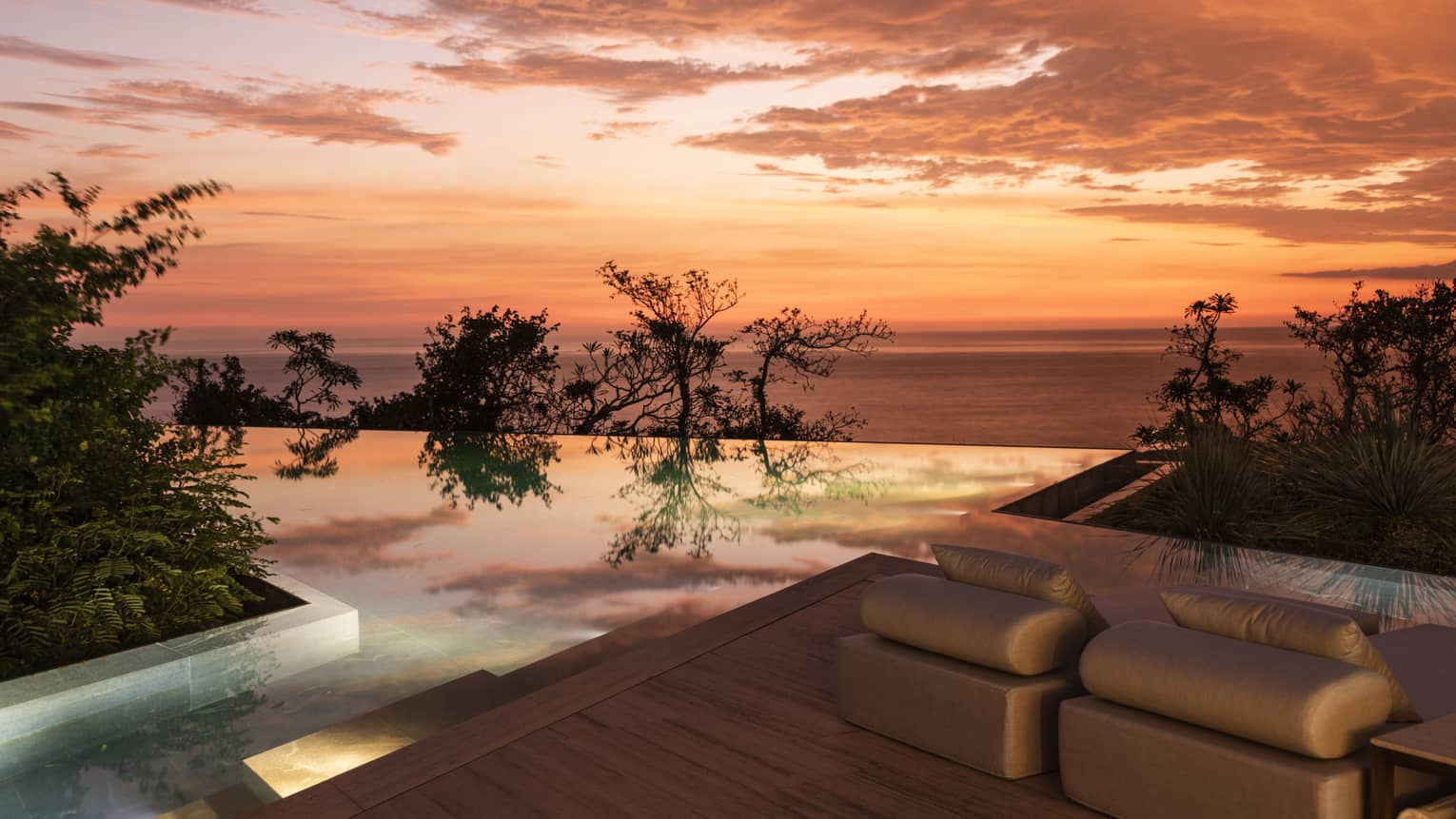 A stunning infinity pool overlooking the ocean at sunset, with soft lounge seating and vibrant orange and pink hues reflecting off the water, surrounded by lush greenery