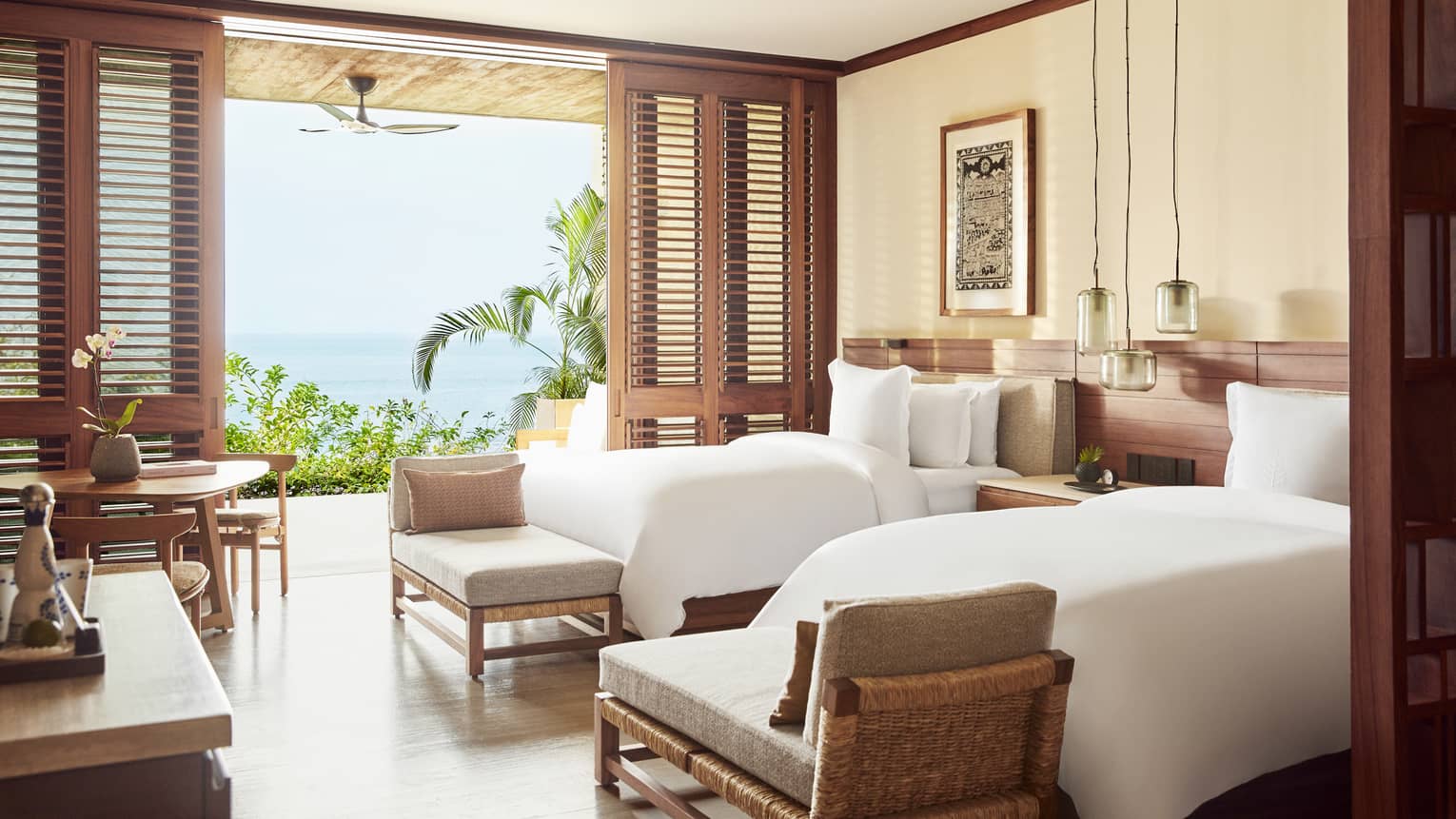 A beachfront room with 2 beds, a small table, benches at the end of each bed and a large open door to outside.
