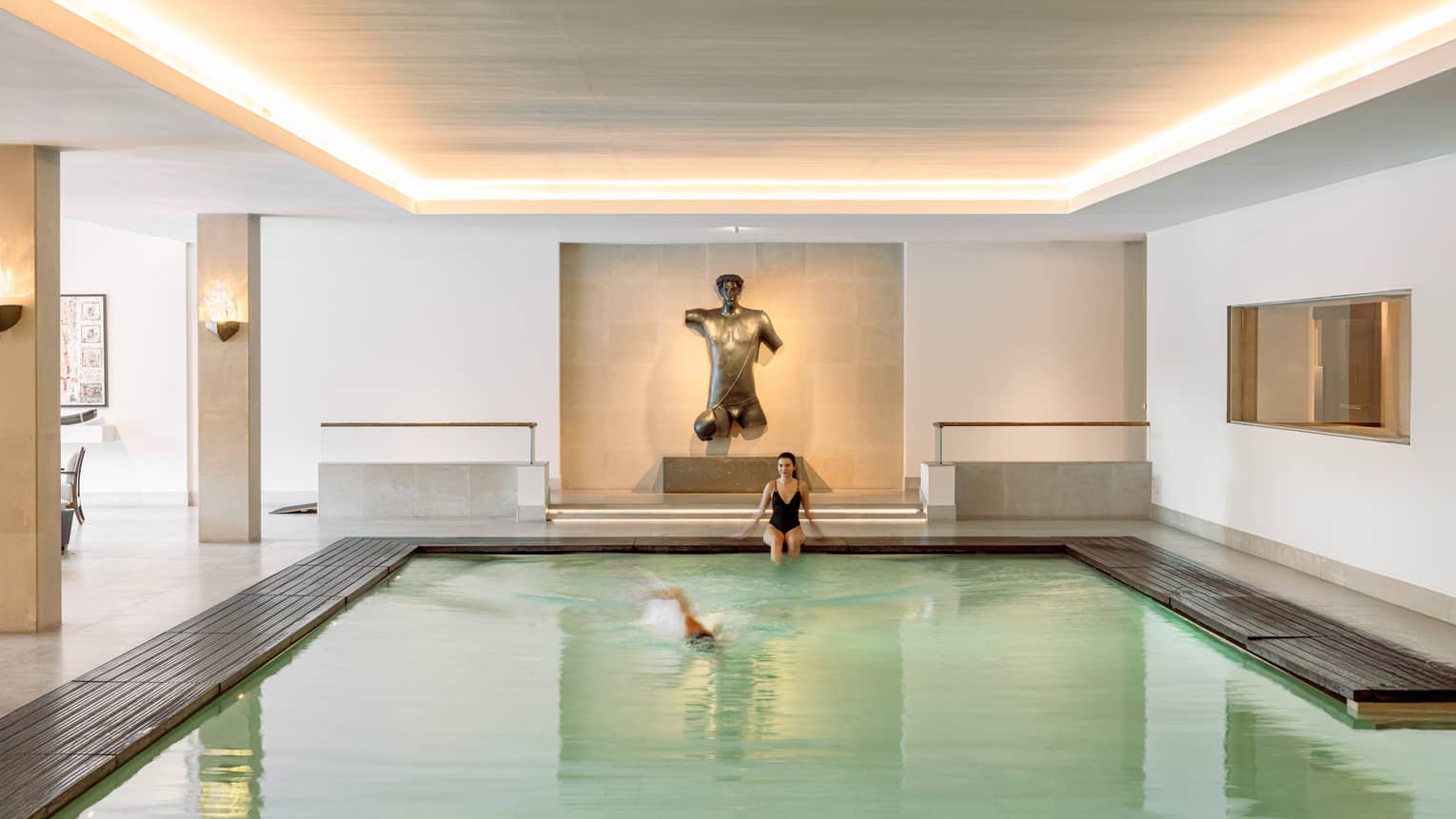 Person sitting at end of indoor lap pool under uplit ceiling while another person swims