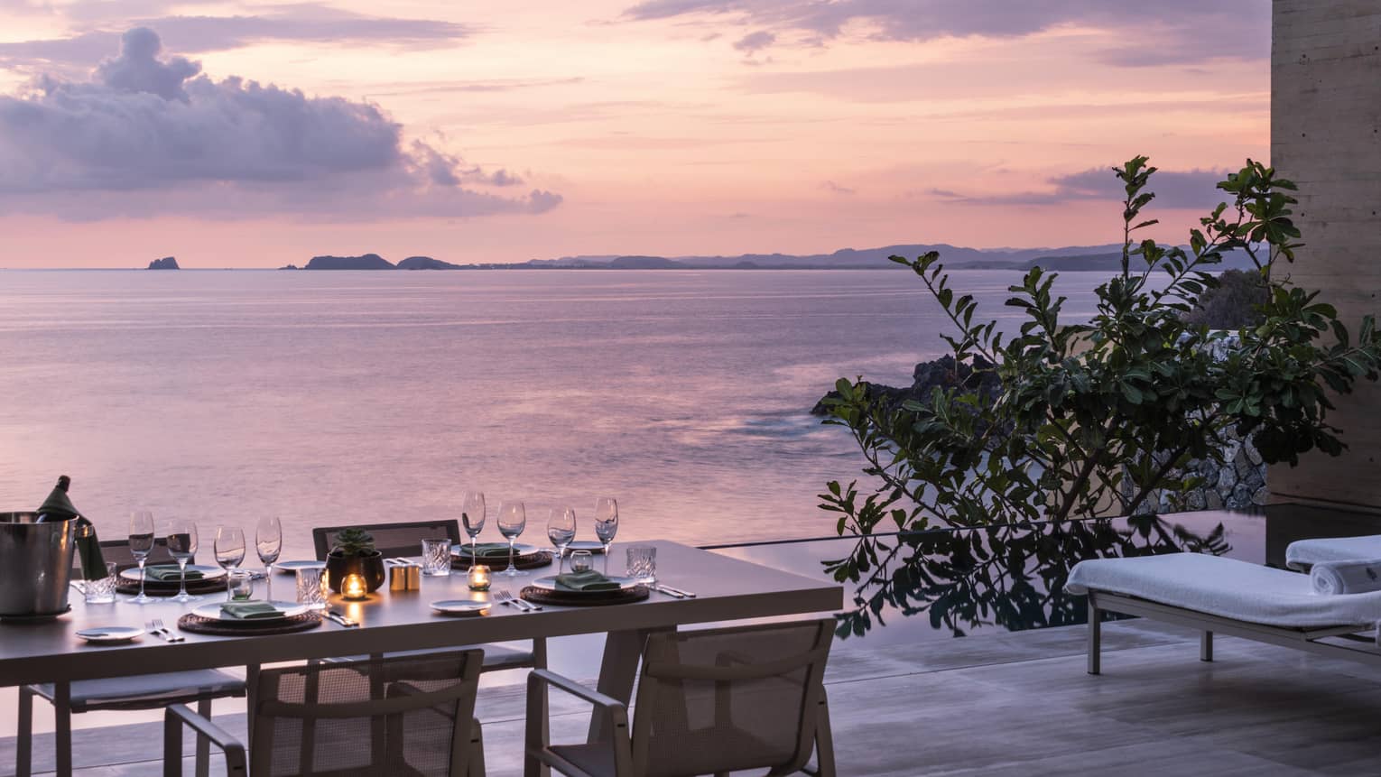 A dining table set for a candlelit meal on a patio overlooking the ocean at sunset. The serene view includes distant islands, soft pink skies and an infinity pool.