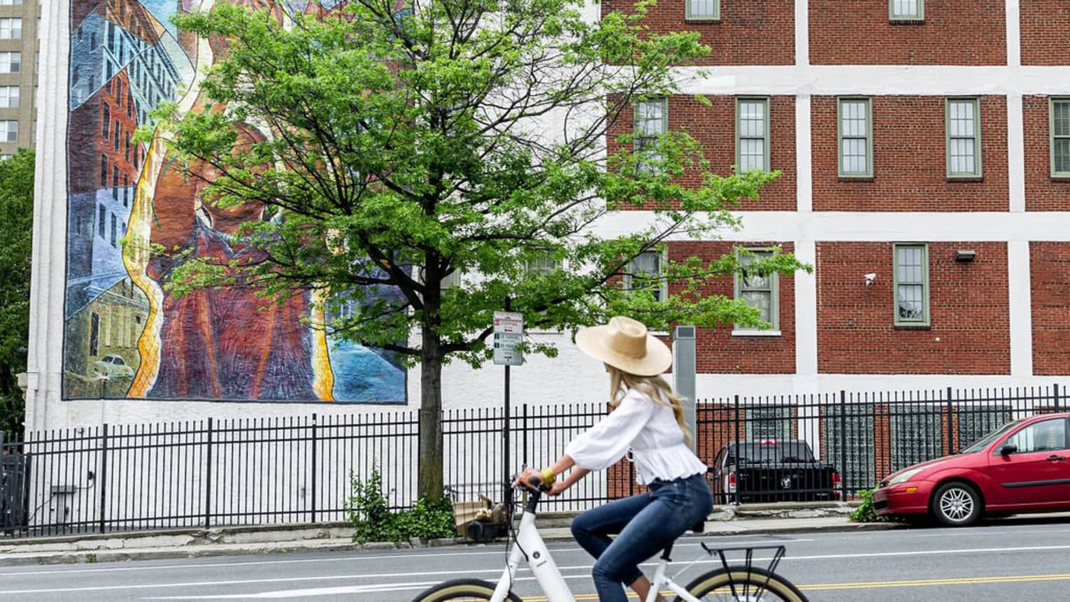 A woman riding a back in a large hat looking at a mural on a white building.