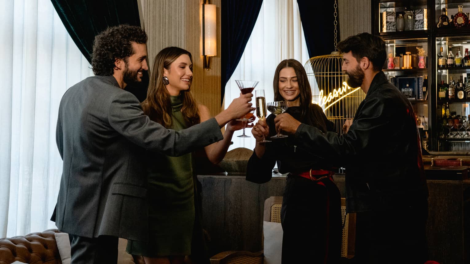 Four people raise their glasses in a toast in a cozy, softly lit lounge setting.