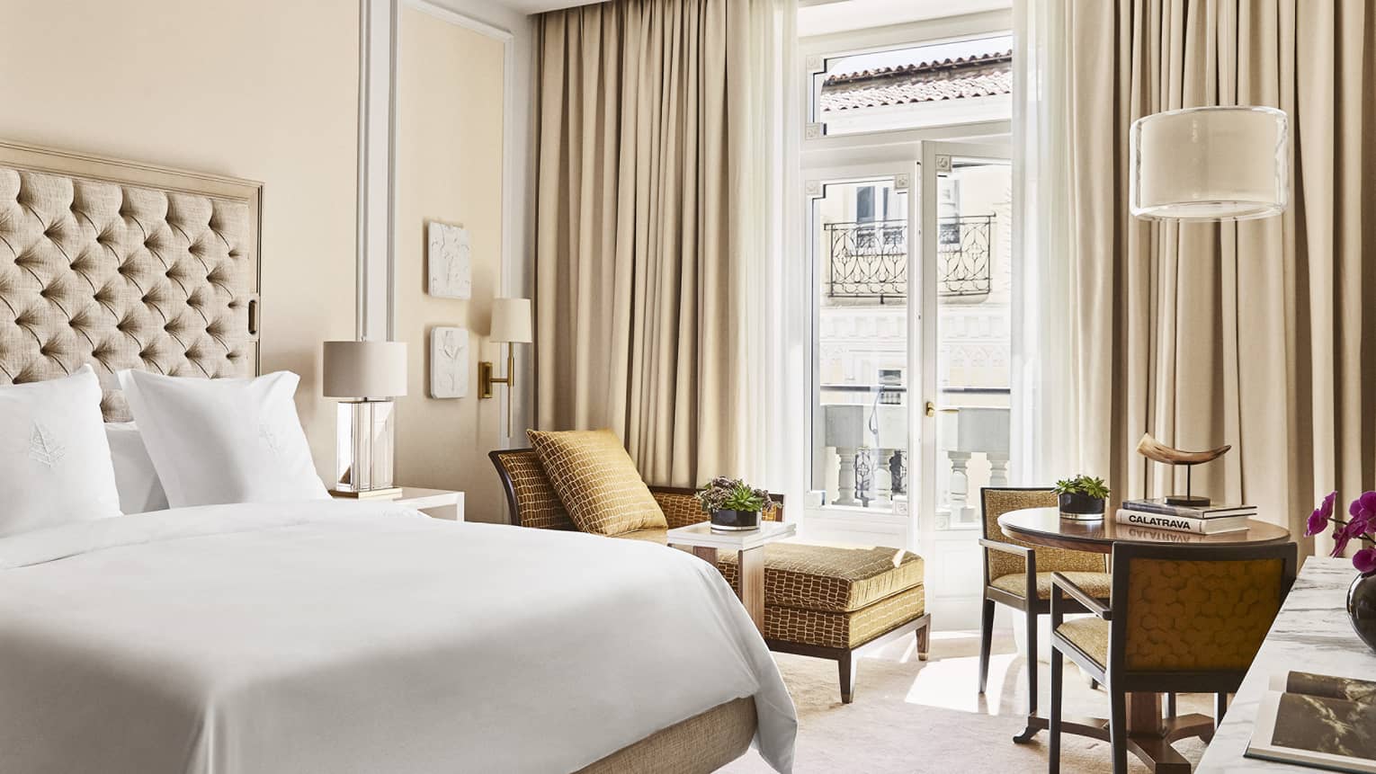Luxury hotel bedroom with king bed, tufted headboard, table with books and horn sculpture, tan chairs and chaise lounge and doors opening to a Juliet balcony
