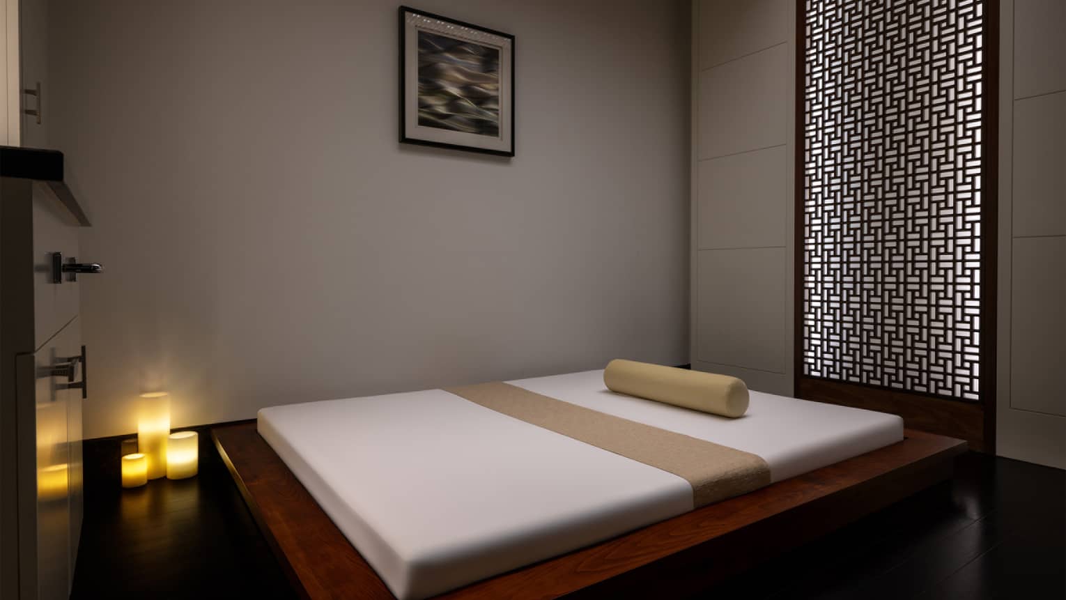 Double-size white floor mat on an elevated wooden platform sits in the centre of a spa treatment room with three candles lit in the corner