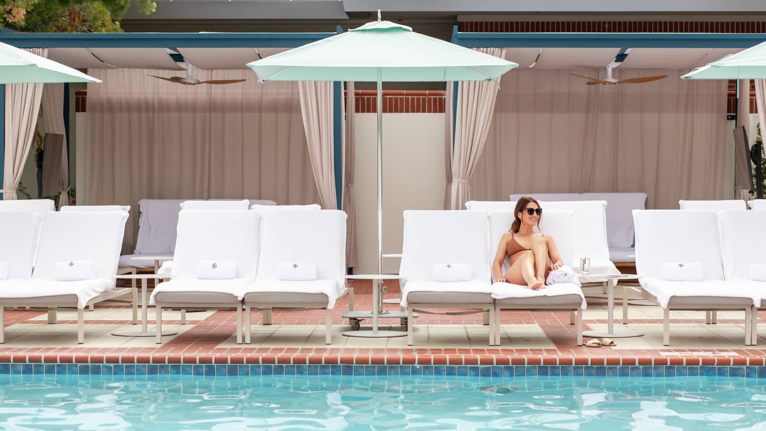 A woman sitting on a lounge chair next to a pool on a sunny day.