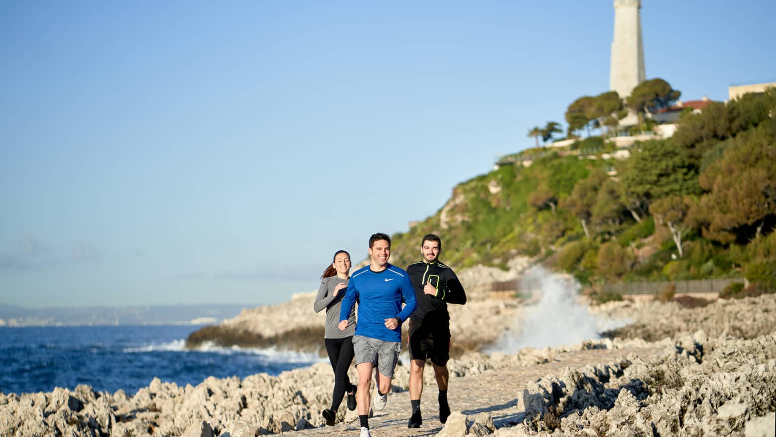 Three people jogging, smiling, on rocky trail beside the sea, lighthouse in backdrop