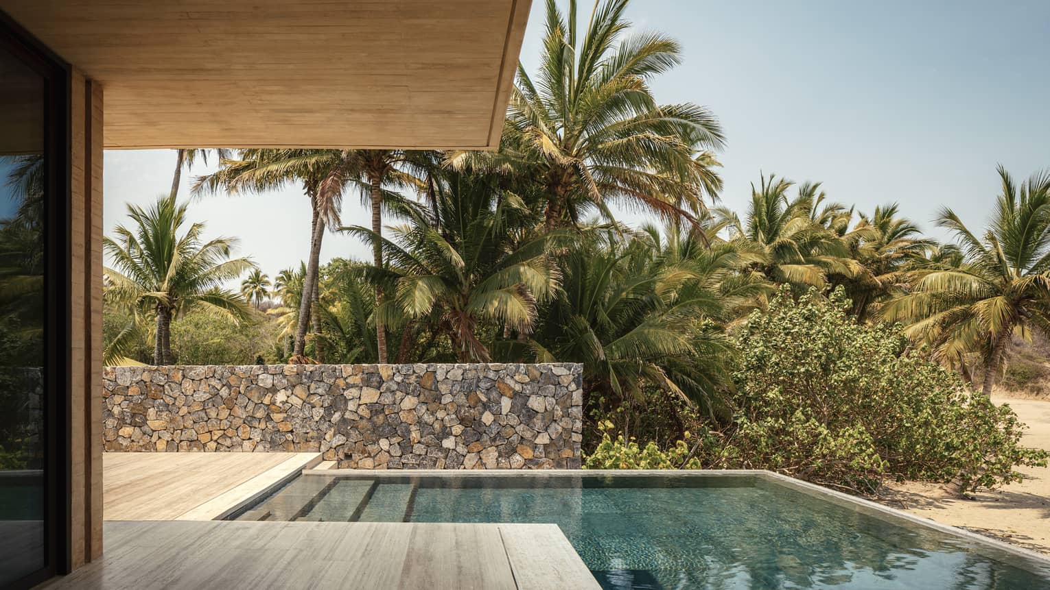 Outdoor poolside area with stone walls, surrounded by lush palm trees and greenery. A covered patio overlooks the tranquil scene under a clear blue sky.
