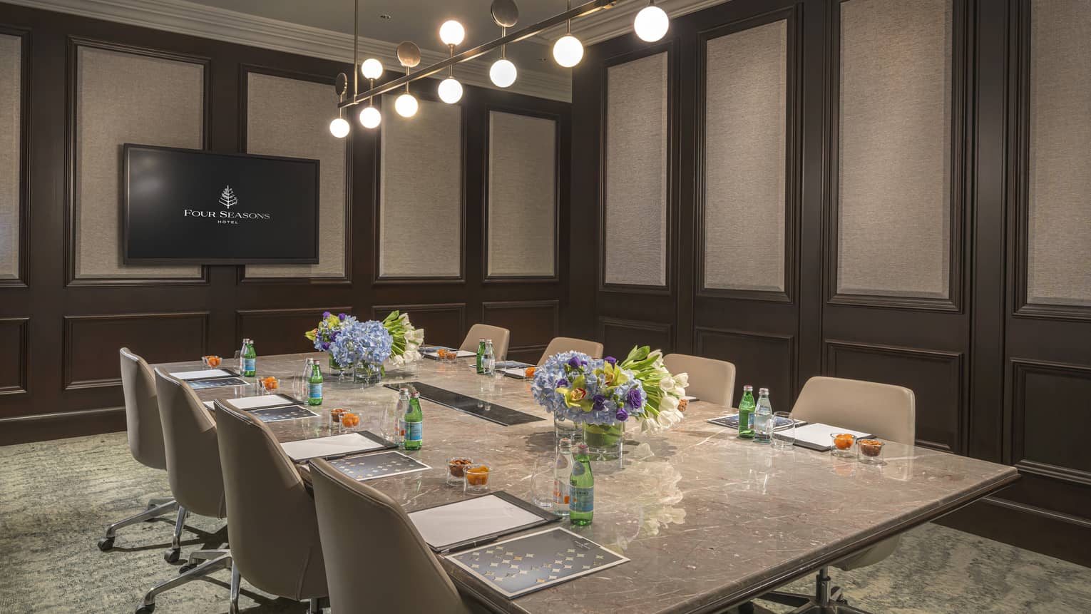 Meeting room with rectangular marble table and four chairs on either side, wall-mounted TV at front of room, green patterned carpet and modern chandelier