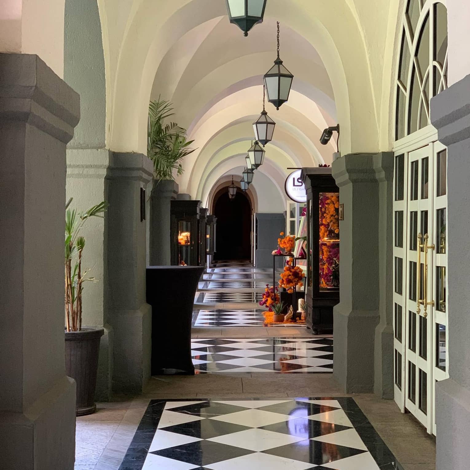 An arched hallway with black and white checker tiles and steel lanterns hanging from the ceiling.