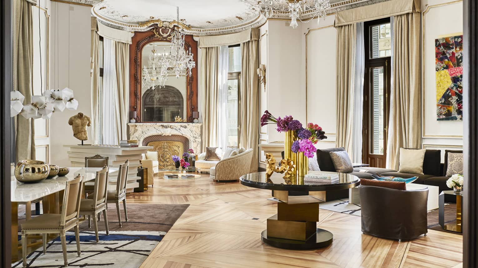 Royal suite living room with high ceilings, ornate chandeliers, grand mirror over a marble fireplace, luxury seating and modern dining table with gold accents