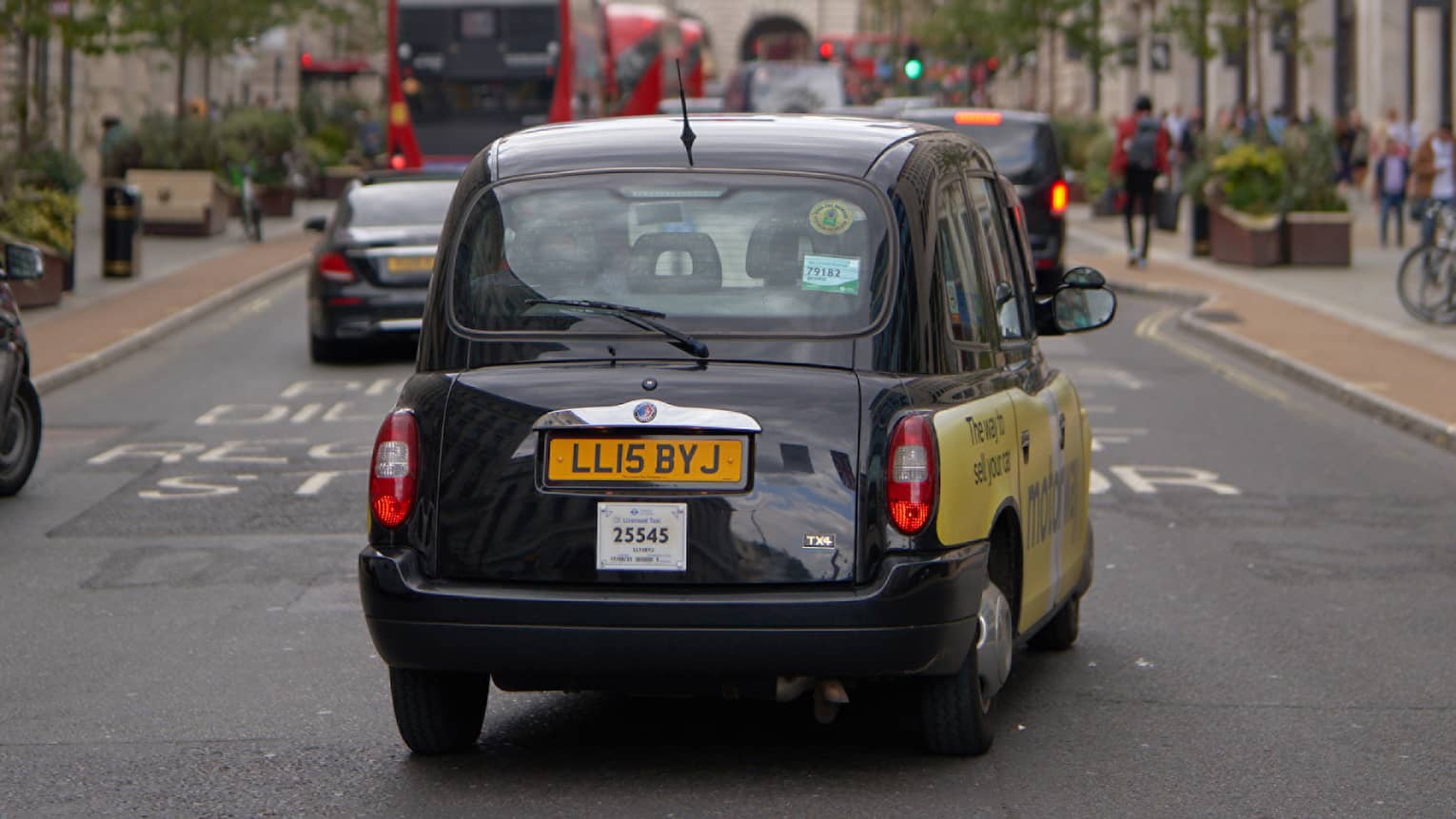 Rear view of London-style black cab, double-decker buses and other cars on road strung across with rows of Union Jack flags.