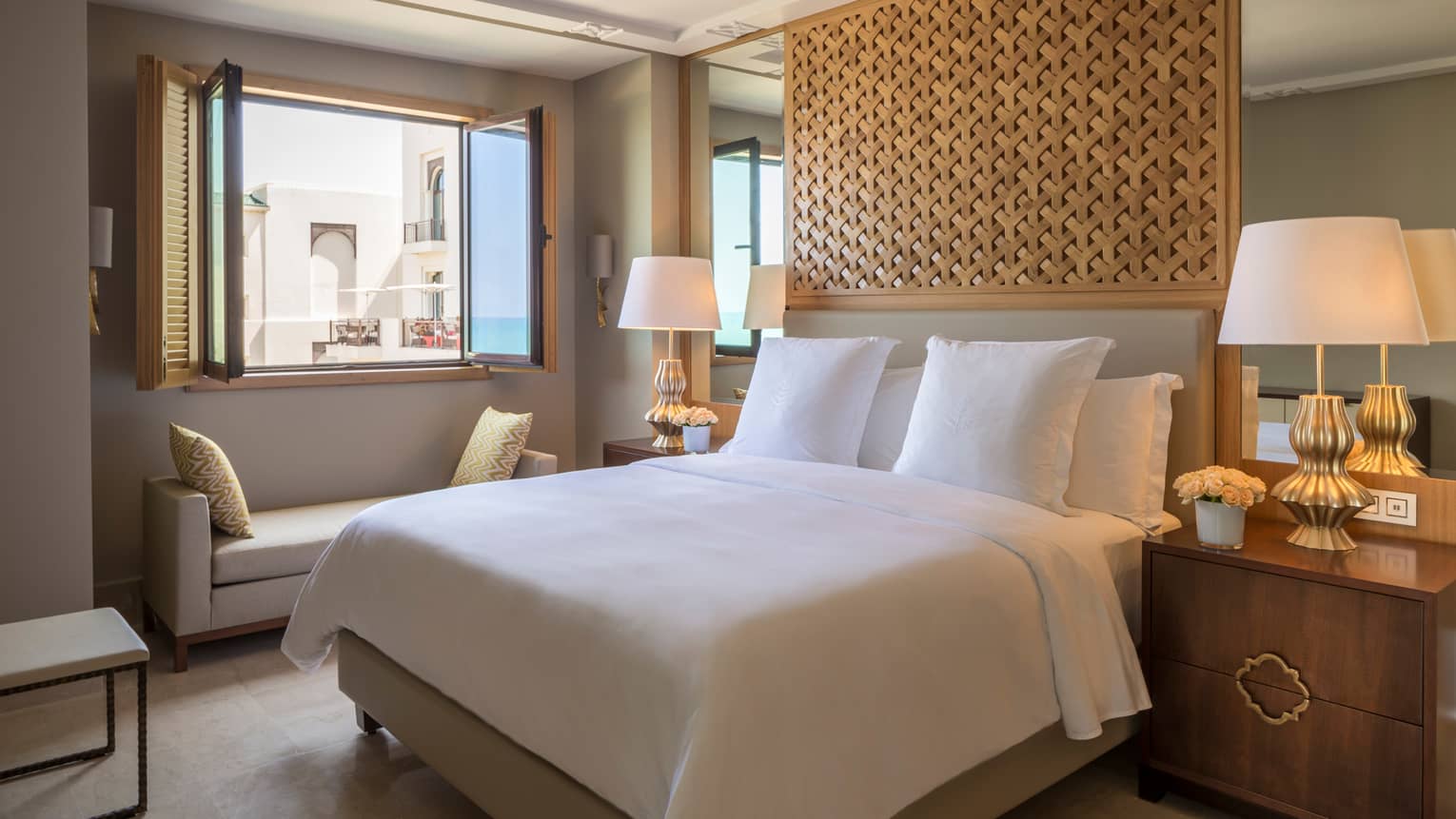 All-white bed with ceiling-high carved wooden headboard flanked by lamps, bench under window