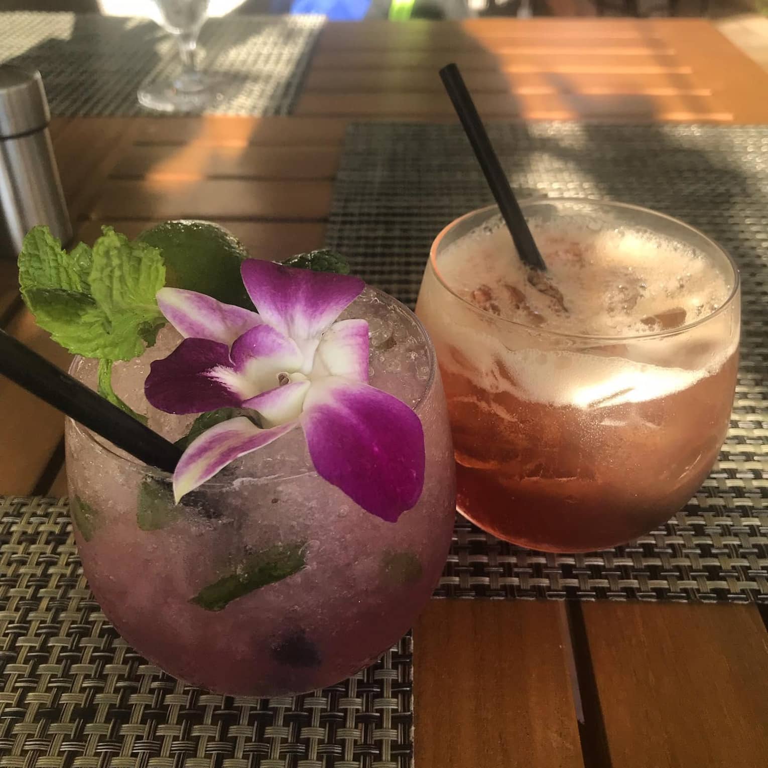 Two refreshing cocktails on a table, one garnished with a purple orchid and mint, and the other with a foamy top