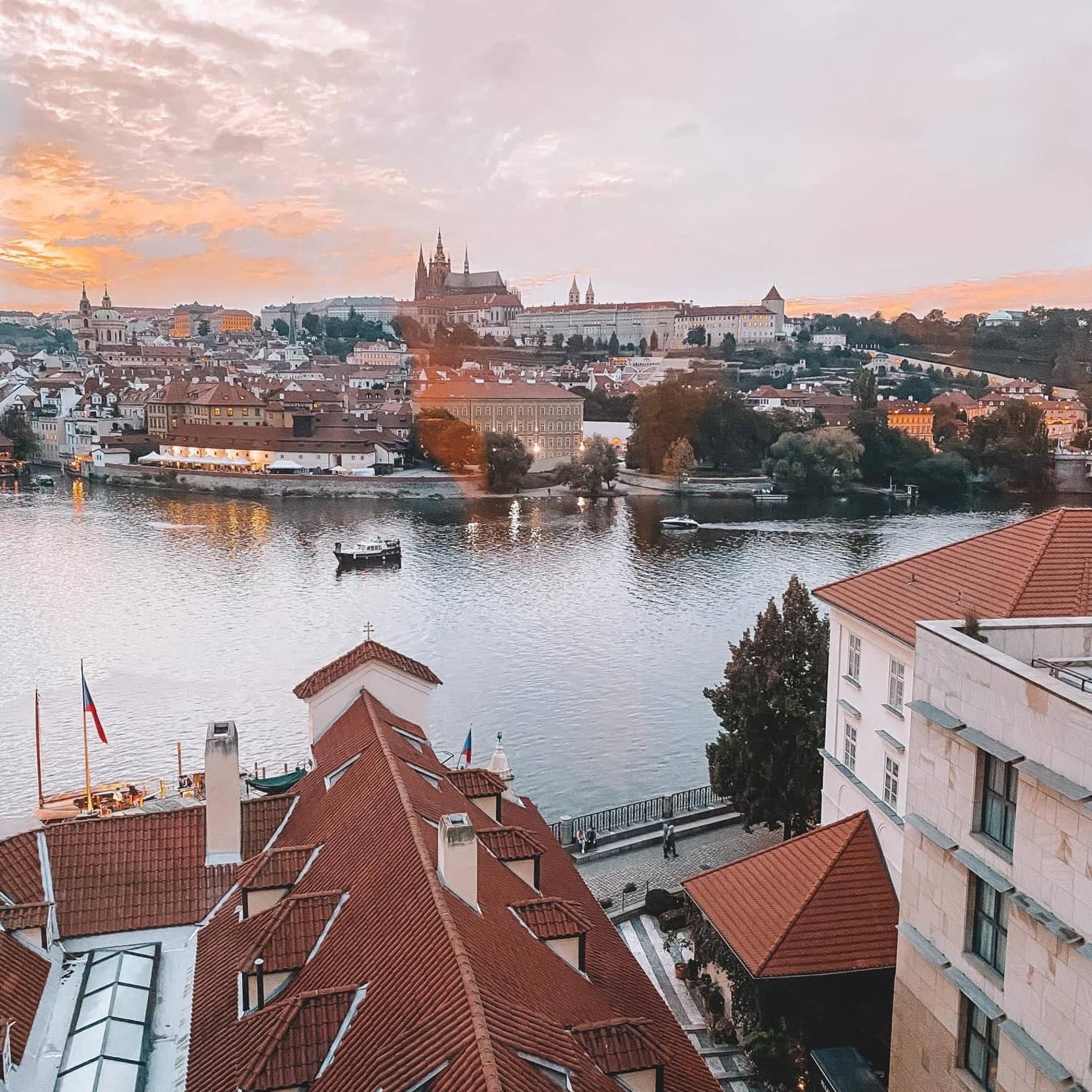 Aerial view of a city with red-roofed buildings along a river, boats on the water and a castle in the background at sunset.