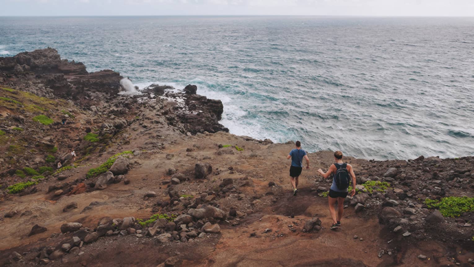 Two people hike down dirt-covered path, rocks towards ocean