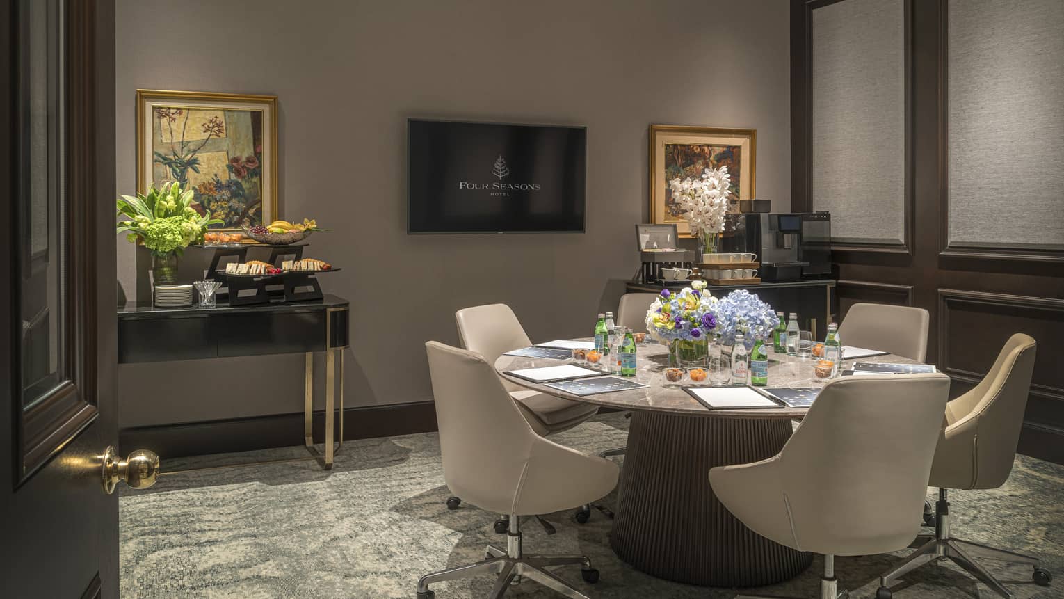 Round marble table with six chairs surrounding, wall-mounted TV at front of room, snack display visible off to the left