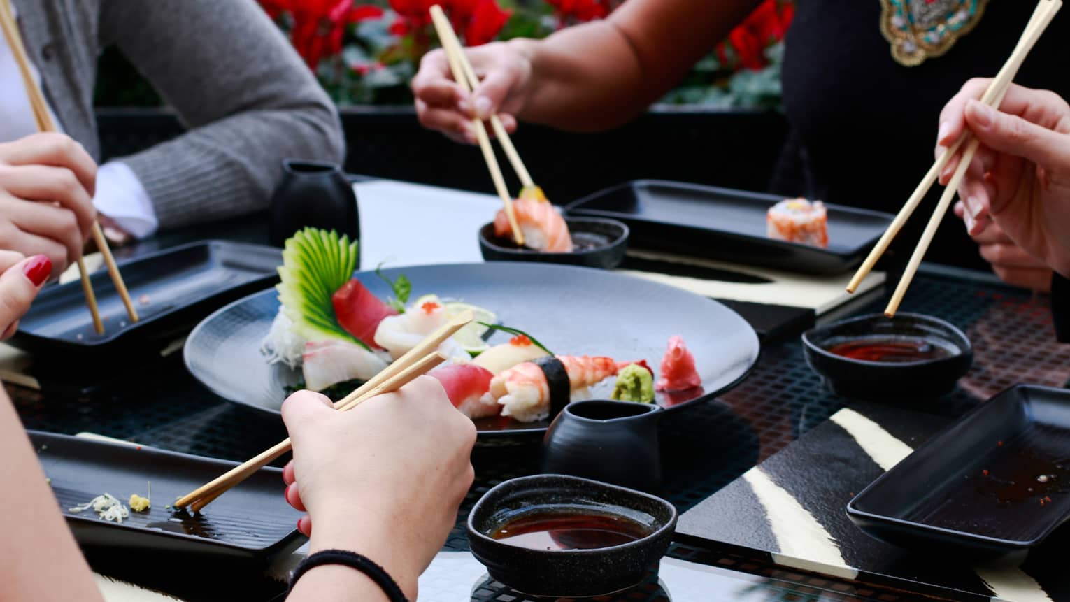 Close-up of four sets of hands holding chopsticks around black plate with sushi rolls, bowls of soy sauce