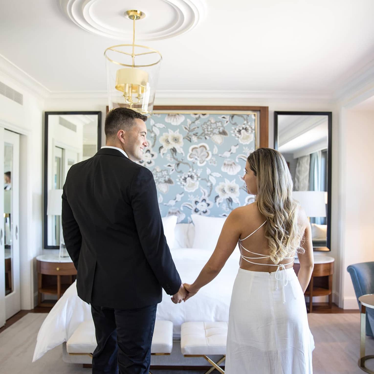 Man wearing suit and woman wearing white dress hold hands in front of hotel bed