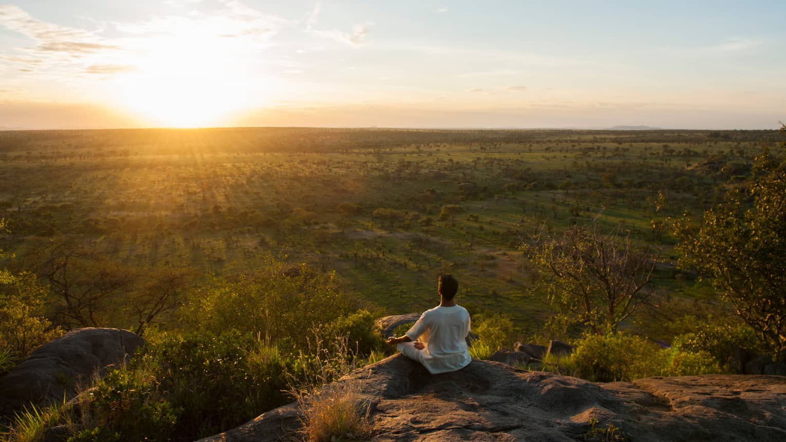 Woman sits crossed-legged, meditates on cliff overlooking Serengeti sunset