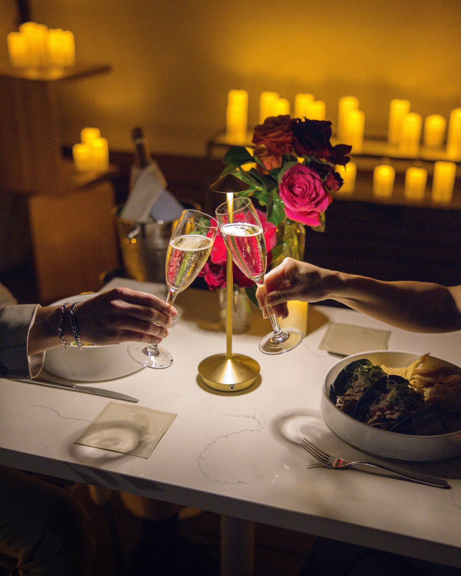 Two hands clink champagne glasses over a dining table set with flowers, candles in the background