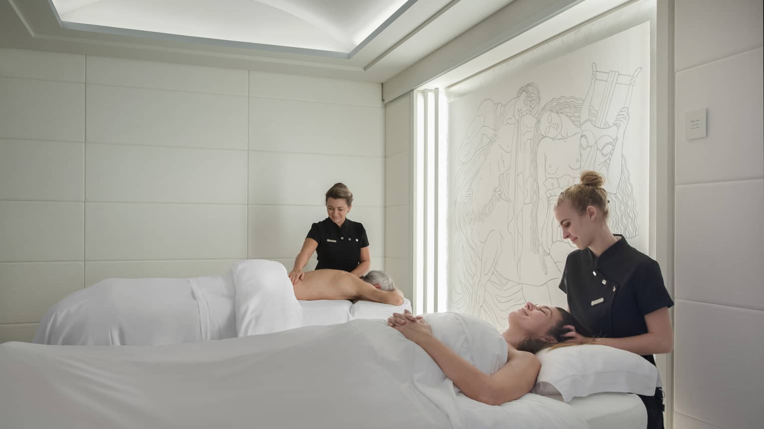 Couple on spa tables under white covers being massaged by spa therapists in couples spa room