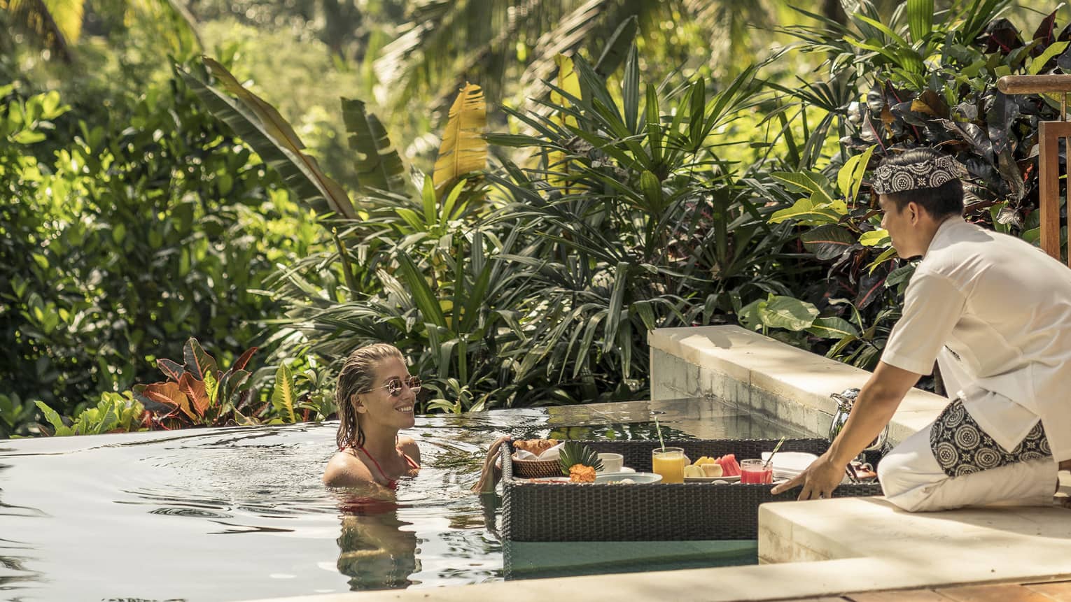 Hotel staff leans over swimming pool, passes floating breakfast tray to woman in water
