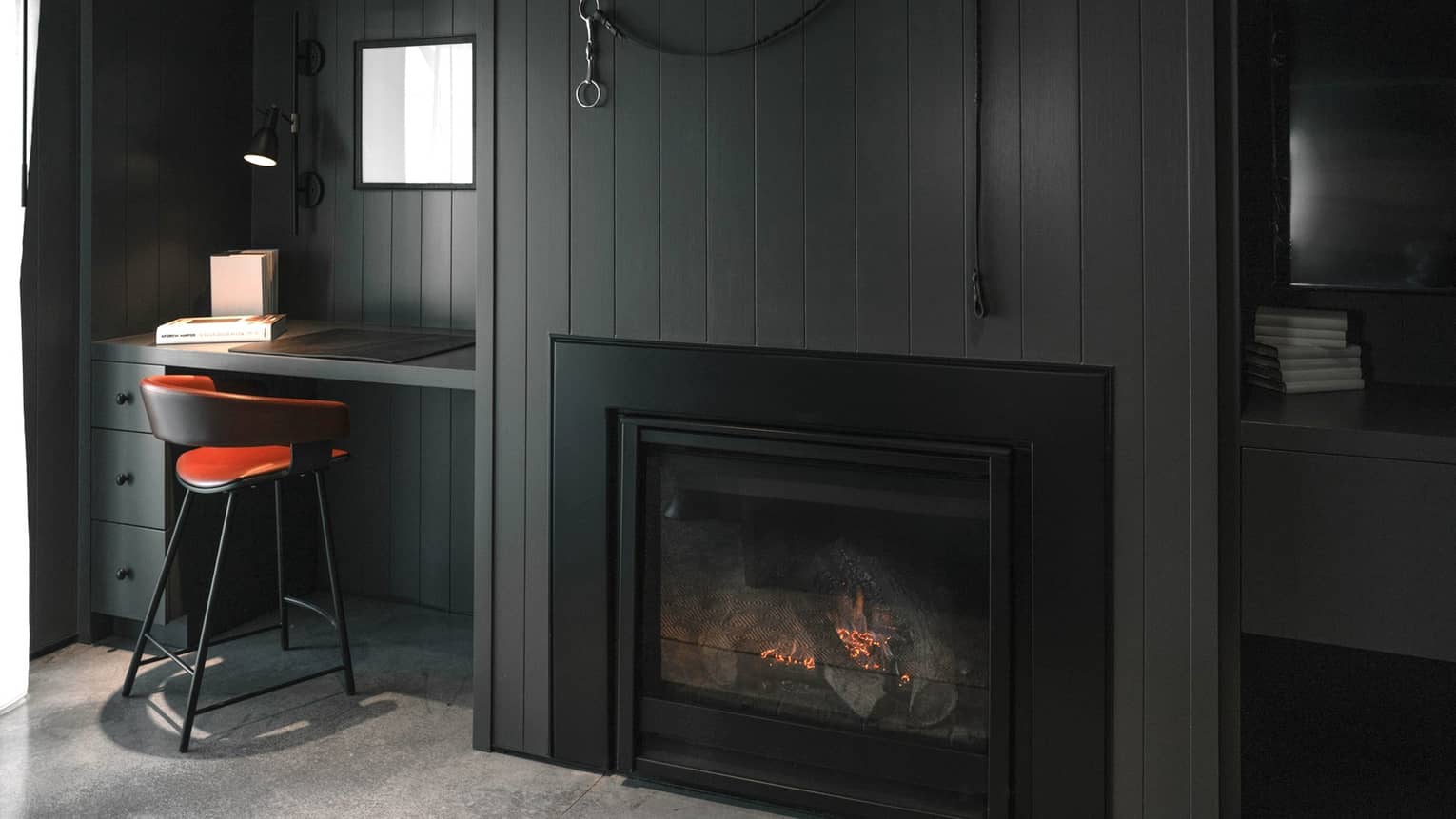 Luxury resort room with dark paneled walls, fireplace, writing desk, orange-accented chair, desk lamp, books and decor on the wall
