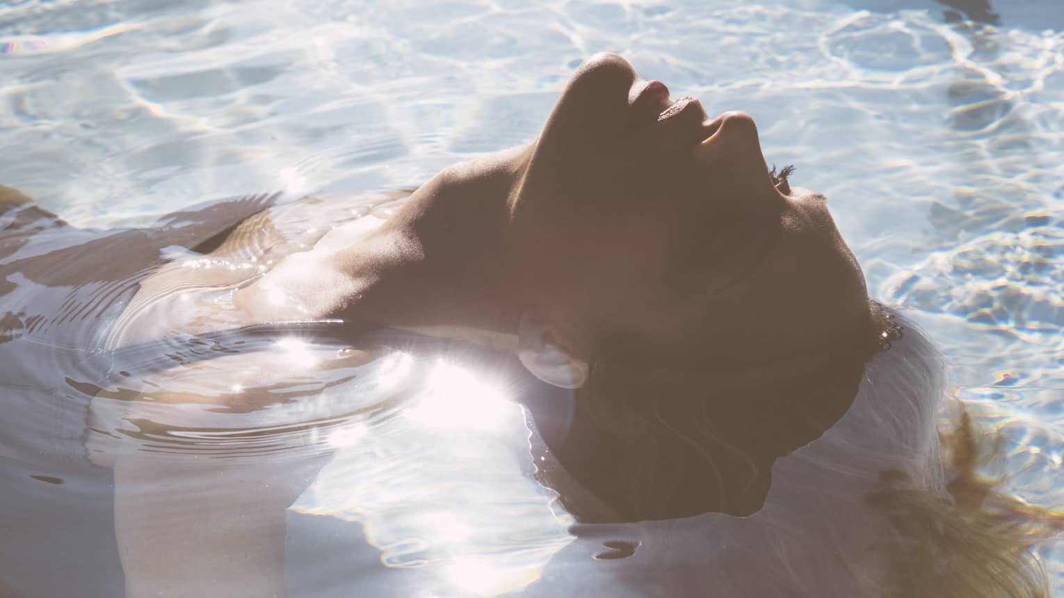 Close-up of woman's head as she floats on her back in pool, sunlight reflecting, her hair flowing in water 
