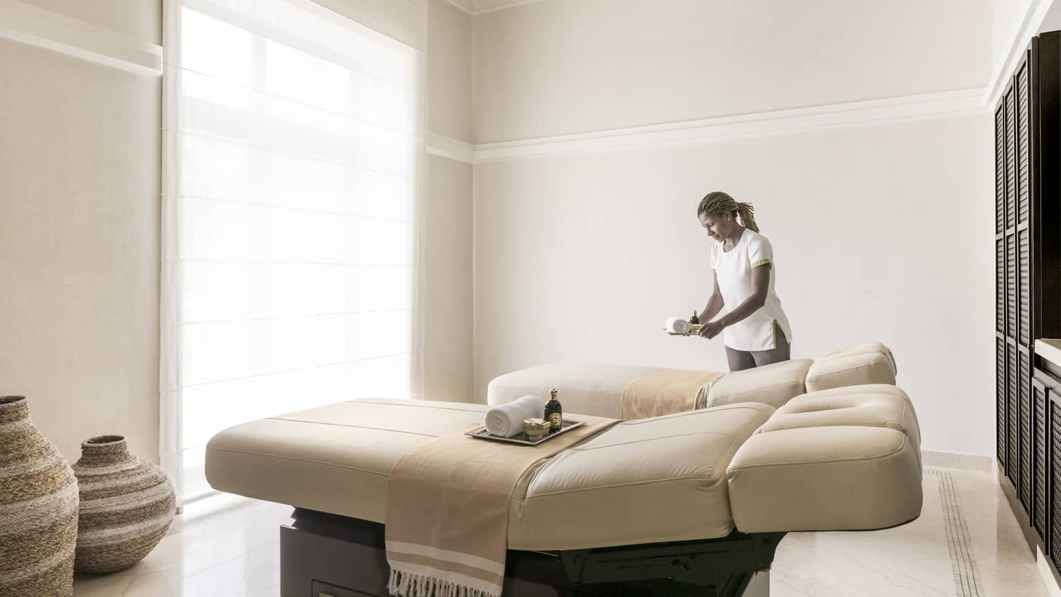 Hotel staff sets tray on Spa Couples Suite massage bed in bright treatment room