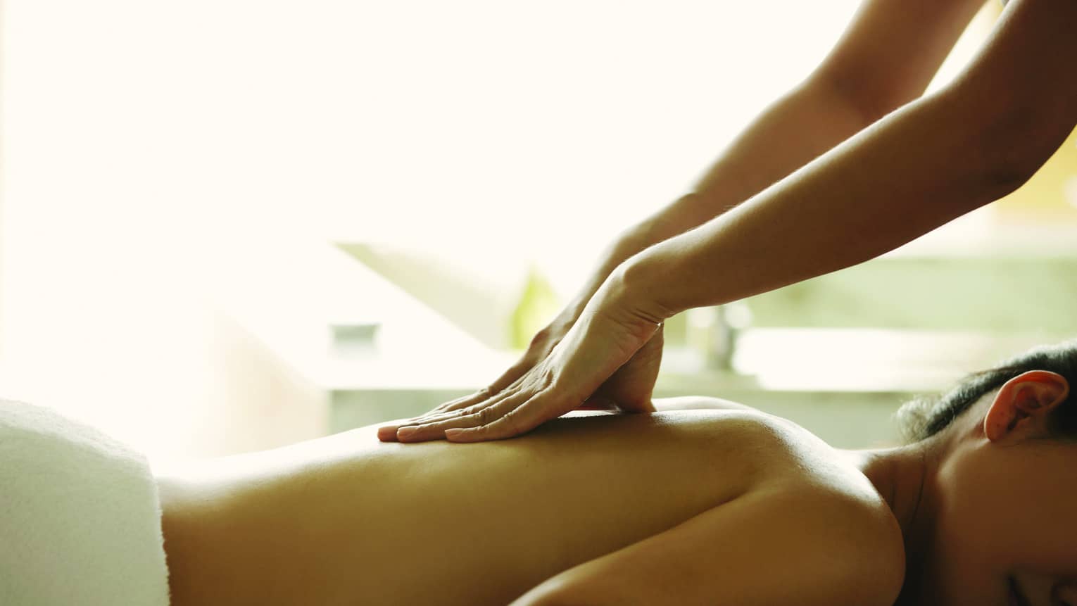 Masseuse reaches over, massages woman's bare back as she lies on spa table